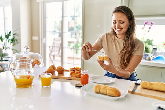 Young beautiful hispanic woman preparing breakfast putting jam on bread at the kitchen