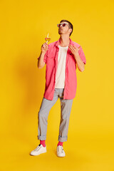 Portrait of young man in pink shirt posing, enjoying wine isolated over vivid yellow background. Summer relaxation