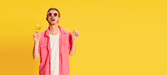 Portrait of young man in pink shirt, funny sunglasses posing with wine glass isolated over vivid yellow background. Relaxed. Flyer