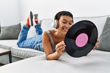 Young hispanic woman listening to music holding vinyl disc lying on sofa at home