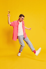 Portrait of young man in pink shirt dancing with wine glass isolated over vivid yellow background. Birthday party