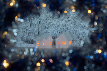 Small white cardboard houses with cut shining windows on dark blue bokeh blurred foreground and snowy trees on background. Shallow depth of focus. Winter decoration set with sparkling lights, models.