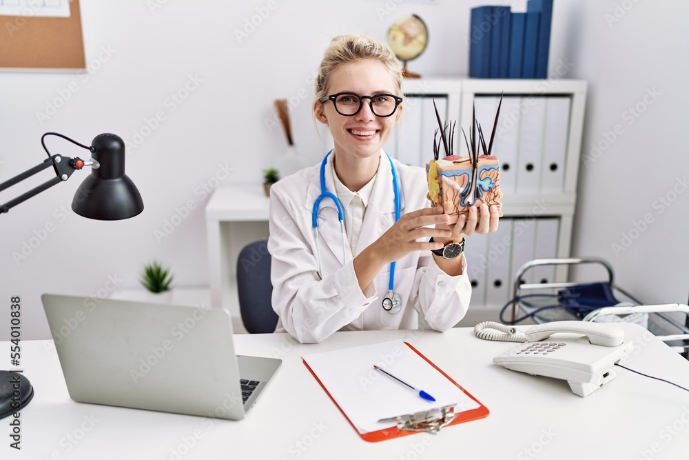 Sticker Young doctor woman holding model of human anatomical skin and hair at the clinic looking positive and happy standing and smiling with a confident smile showing teeth