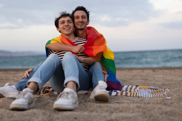 Beautiful gay young couple embraces and holds a rainbow flag. Happy couple enjoy at the beach.