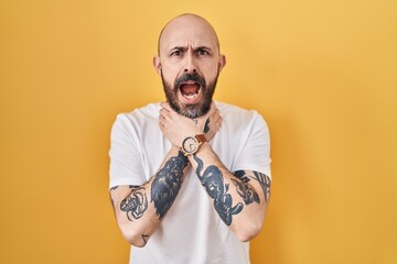 Young hispanic man with tattoos standing over yellow background shouting suffocate because painful strangle. health problem. asphyxiate and suicide concept.