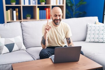 Hispanic man with tattoos using laptop at home smiling with an idea or question pointing finger with happy face, number one