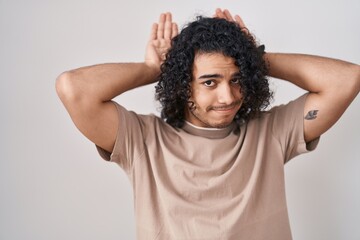 Hispanic man with curly hair standing over white background doing bunny ears gesture with hands palms looking cynical and skeptical. easter rabbit concept.