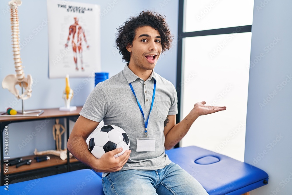 Wall mural hispanic man with curly hair working as football physiotherapist pointing aside with hands open palm