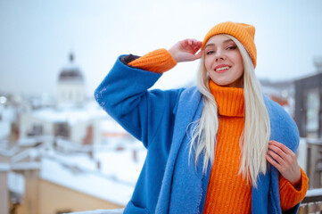 Happy smiling fashionable woman wearing trendy blue winter coat,  woolen scarf, orange knitted beanie hat, turtleneck sweater, posing outdoor. Copy, empty space for text