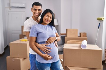 Young latin couple expecting baby hugging each other standing at new home - obrazy, fototapety, plakaty