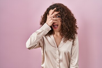 Hispanic woman with curly hair standing over pink background peeking in shock covering face and eyes with hand, looking through fingers with embarrassed expression.