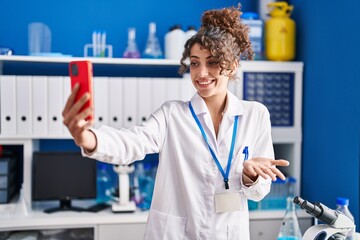 Hispanic woman with curly hair working at scientist laboratory doing selfie celebrating achievement...
