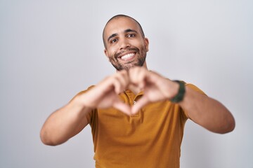 Hispanic man with beard standing over white background smiling in love doing heart symbol shape with hands. romantic concept.