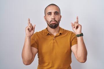 Hispanic man with beard standing over white background pointing up looking sad and upset,...