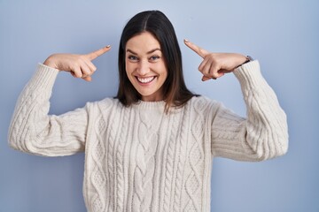 Young brunette woman standing over blue background smiling pointing to head with both hands finger, great idea or thought, good memory