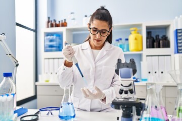 Young hispanic woman wearing scientist uniform using pipette working at laboratory