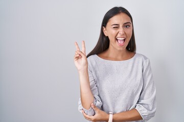 Young hispanic woman standing over white background smiling with happy face winking at the camera...