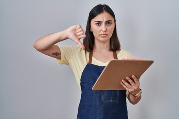 Hispanic girl wearing apron holding clipboard with angry face, negative sign showing dislike with...