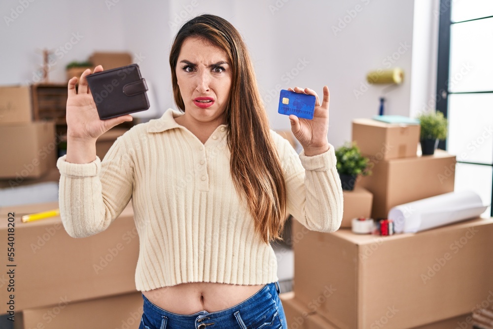 Sticker young hispanic woman moving to a new home holding wallet and credit card clueless and confused expre