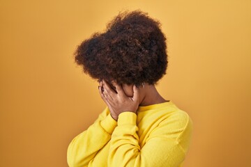 Young african american woman standing over yellow background with sad expression covering face with...