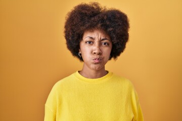Obraz na płótnie Canvas Young african american woman standing over yellow background puffing cheeks with funny face. mouth inflated with air, crazy expression.