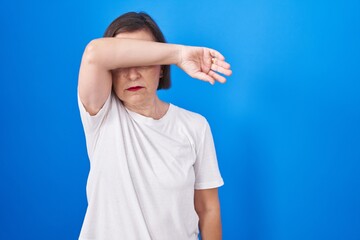 Middle age hispanic woman standing over blue background covering eyes with arm, looking serious and sad. sightless, hiding and rejection concept
