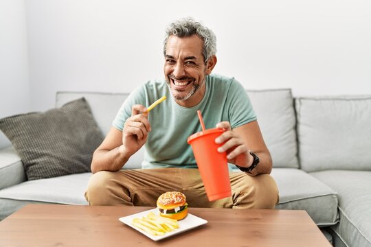 Middle age grey-haired man eating fried potatoes sitting on sofa at home