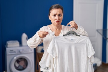 Middle age hispanic woman holding shirt on hanger and detergent powder skeptic and nervous,...