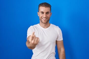 Fototapeta premium Young caucasian man standing over blue background beckoning come here gesture with hand inviting welcoming happy and smiling