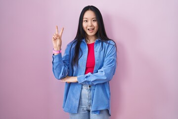 Young asian woman standing over pink background smiling with happy face winking at the camera doing victory sign with fingers. number two.