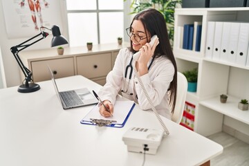 Young beautiful hispanic woman doctor talking on telephone writing on document at clinic