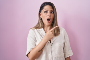 Blonde woman standing over pink background surprised pointing with finger to the side, open mouth amazed expression.