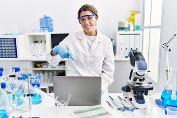 Young hispanic woman wearing scientist uniform working at laboratory smiling happy pointing with...