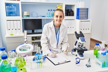 Young blonde woman wearing scientist uniform holding test tubes writing on checklist at laboratory