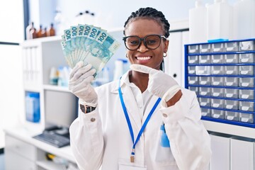African woman with braids working at scientist laboratory holding money smiling happy pointing with hand and finger