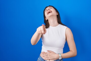 Young caucasian woman standing over blue background laughing at you, pointing finger to the camera...