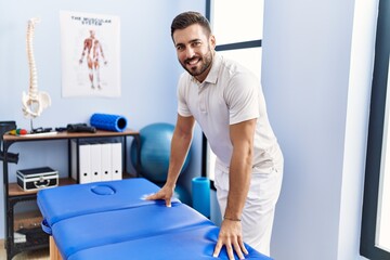 Young hispanic man wearing physiotherapist uniform standing at clinic