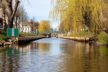 View from the canal in Friedrichsdorf on the North Sea to an old arched bridge with houses, green area and hanging willow tree