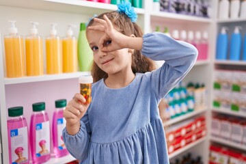 Young little girl holding pills at the pharmacy smiling happy doing ok sign with hand on eye looking through fingers