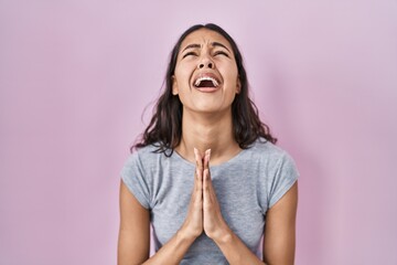 Young brazilian woman wearing casual t shirt over pink background begging and praying with hands...