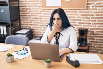Young girl with blue hair working at the office doing video call with laptop covering mouth with...