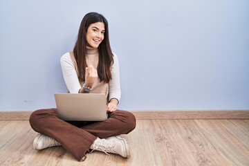 Young brunette woman working using computer laptop sitting on the floor beckoning come here gesture with hand inviting welcoming happy and smiling