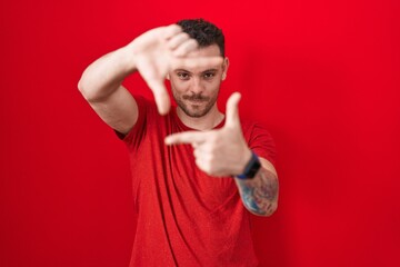 Young hispanic man standing over red background smiling making frame with hands and fingers with happy face. creativity and photography concept.