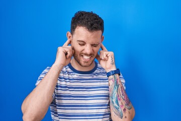 Young hispanic man standing over blue background covering ears with fingers with annoyed expression for the noise of loud music. deaf concept.