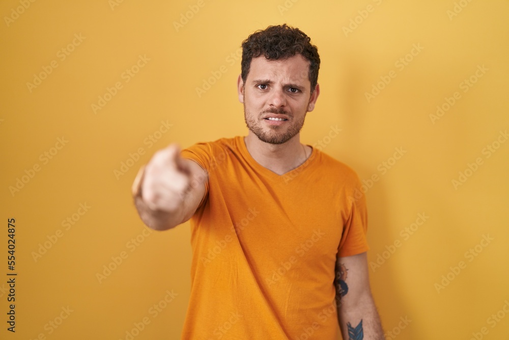 Sticker young hispanic man standing over yellow background pointing displeased and frustrated to the camera,