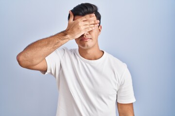 Hispanic man standing over blue background covering eyes with hand, looking serious and sad....