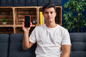 Hispanic man holding smartphone showing blank screen thinking attitude and sober expression looking self confident