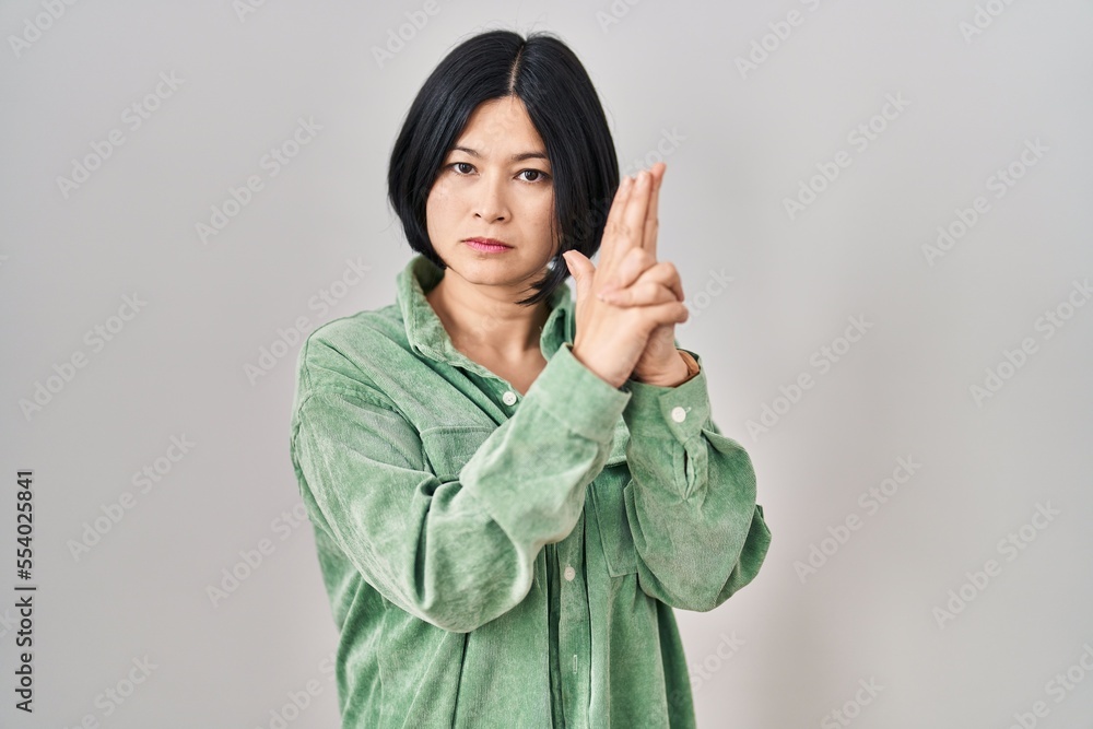 Sticker Young asian woman standing over white background holding symbolic gun with hand gesture, playing killing shooting weapons, angry face