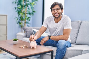 Young hispanic man inserting coin on piggy bank at home