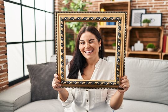 Young hispanic woman holding empty frame winking looking at the camera with sexy expression, cheerful and happy face.
